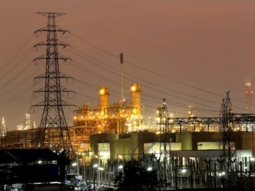 Upplyst kraftverk vid vatten med elmaster i kvällsljus