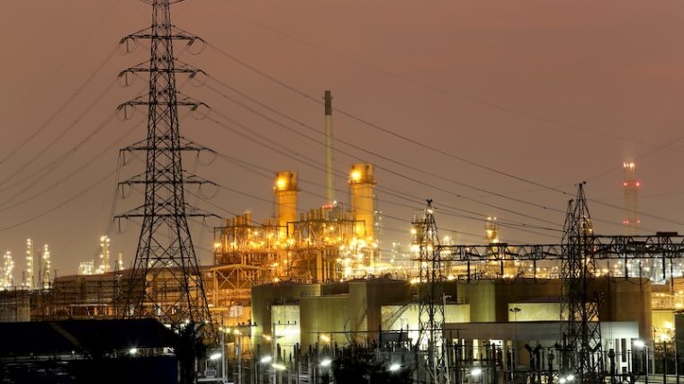 Upplyst kraftverk vid vatten med elmaster i kvällsljus