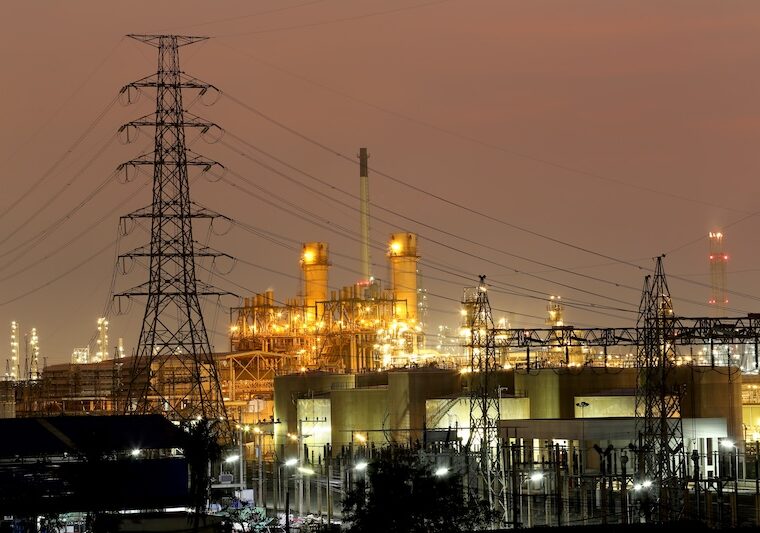 Upplyst kraftverk vid vatten med elmaster i kvällsljus